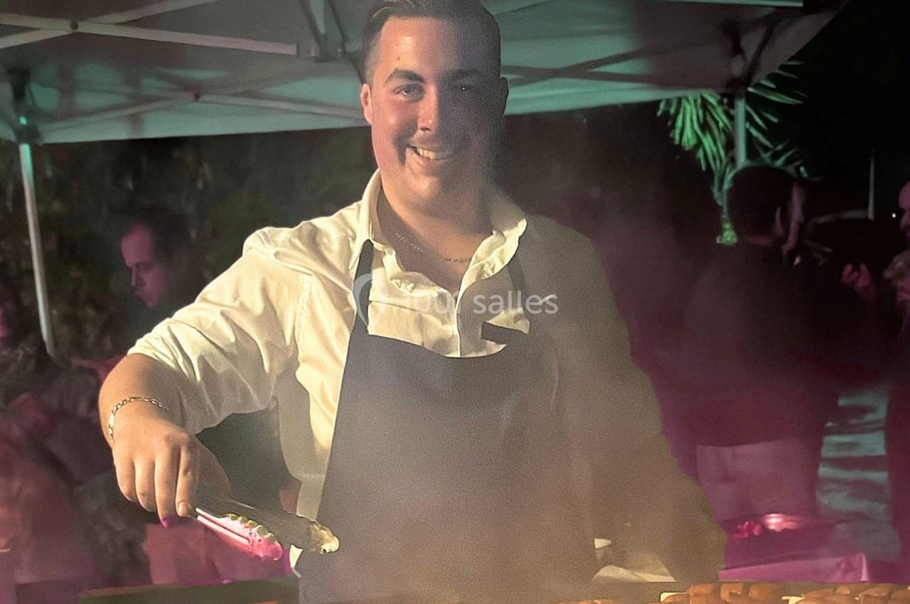 Un homme en tablier cuisine des aliments sur un gril en extérieur, sous un éclairage nocturne.