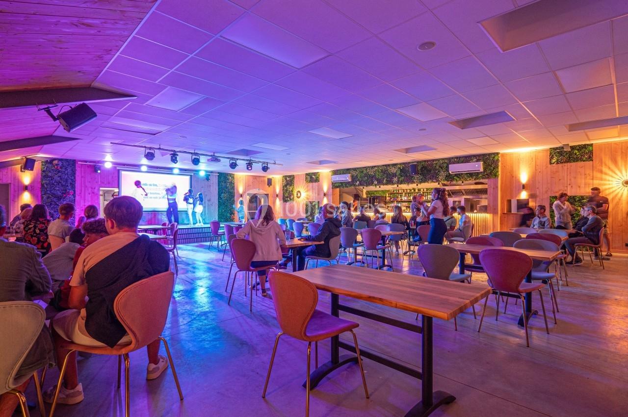 Salle lumineuse avec des tables et des chaises, des personnes assises et debout, et une scène éclairée en arrière-plan.