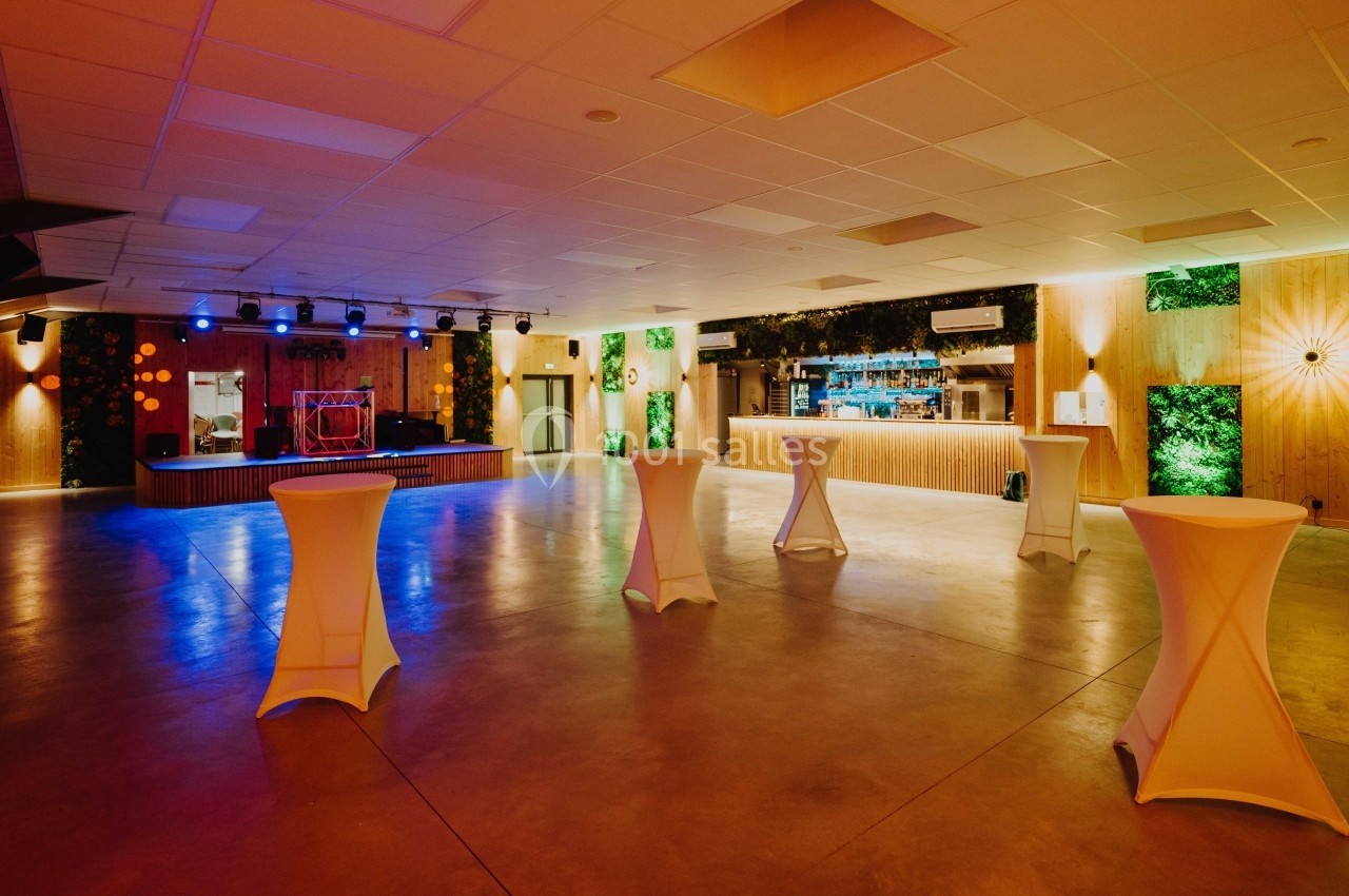 Salle de réception spacieuse avec éclairage tamisé, tables hautes et scène équipée de lumières colorées.