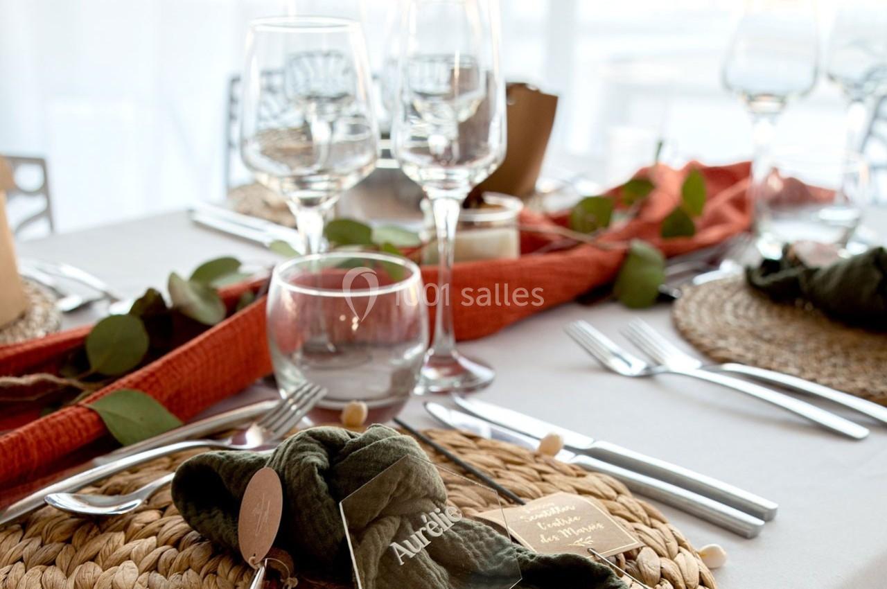 Table dressée avec des assiettes sur sets en osier, verres, couverts, serviettes vertes et décoration végétale.