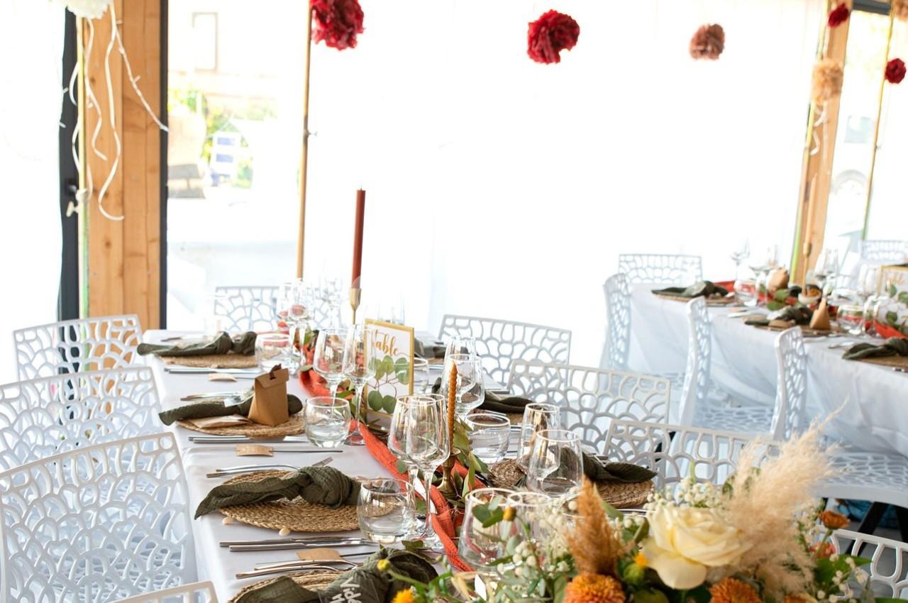 Tables décorées pour un repas festif, avec nappes blanches, fleurs colorées et serviettes pliées sur des assiettes.