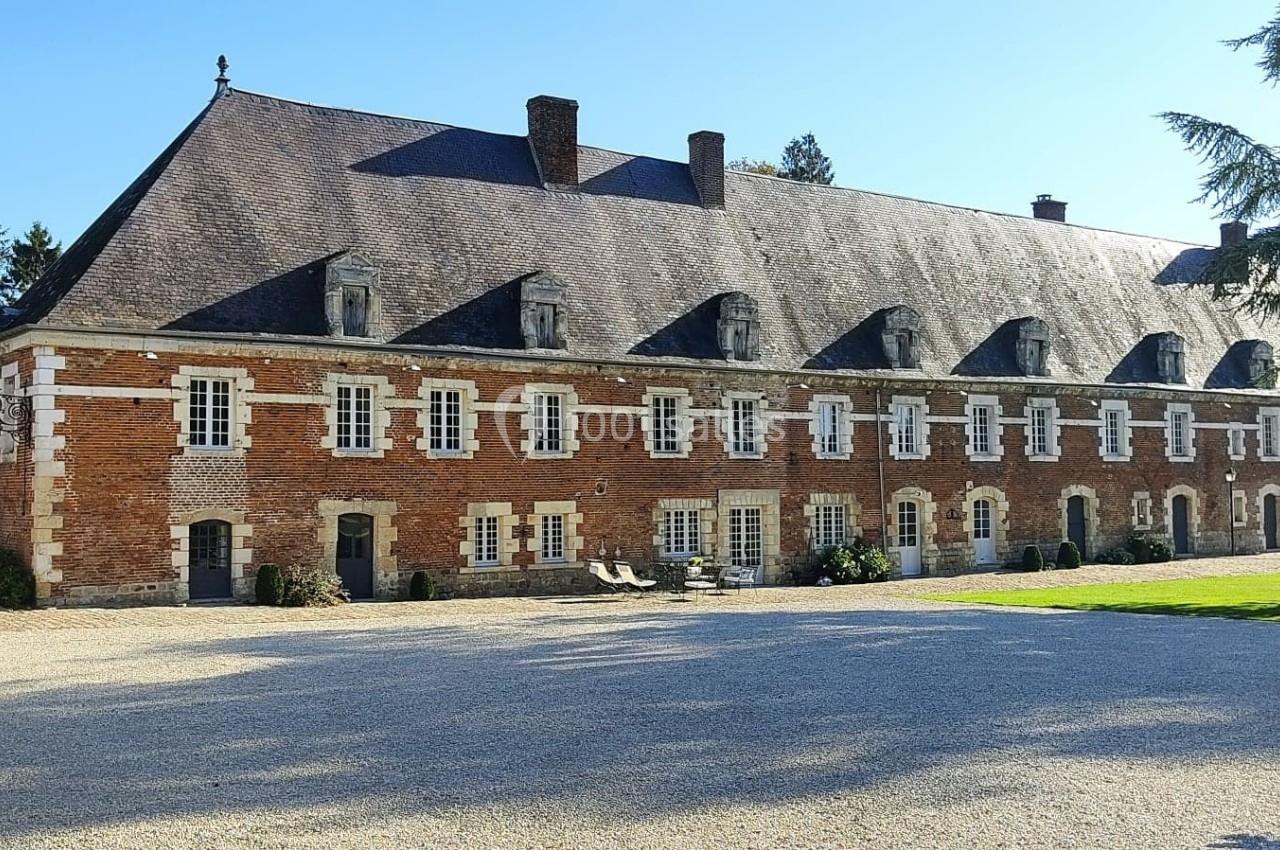 Bâtiment historique en briques rouges avec toit en ardoise, entouré d'une cour gravillonnée et d'espaces verts.