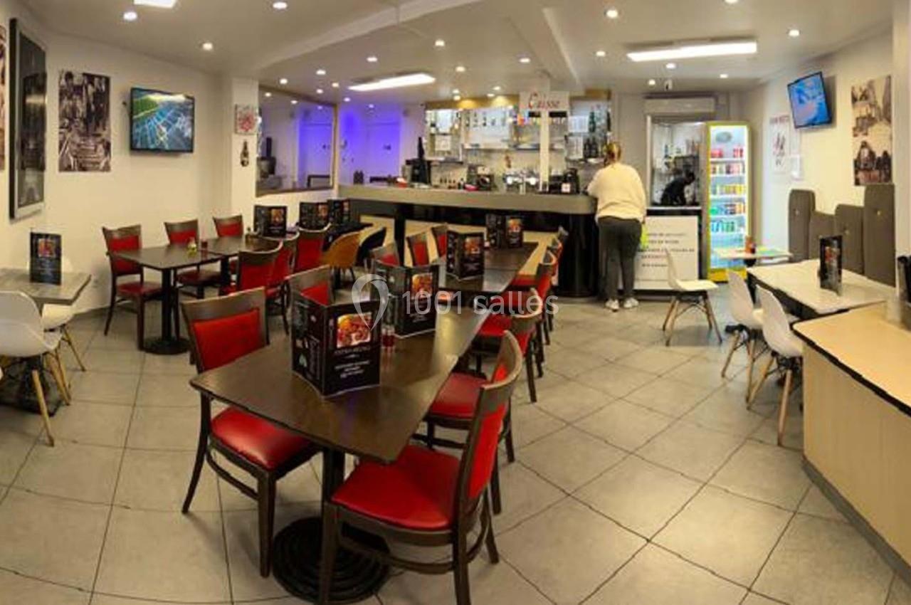 Salle de restaurant avec tables et chaises rouges et noires, comptoir au fond et une personne près d'un réfrigérateur.
