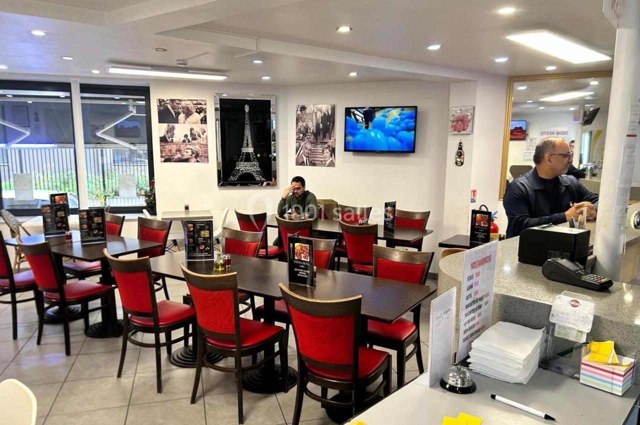 Salle de restaurant lumineuse avec tables et chaises rouges, un client assis et un employé au comptoir.
