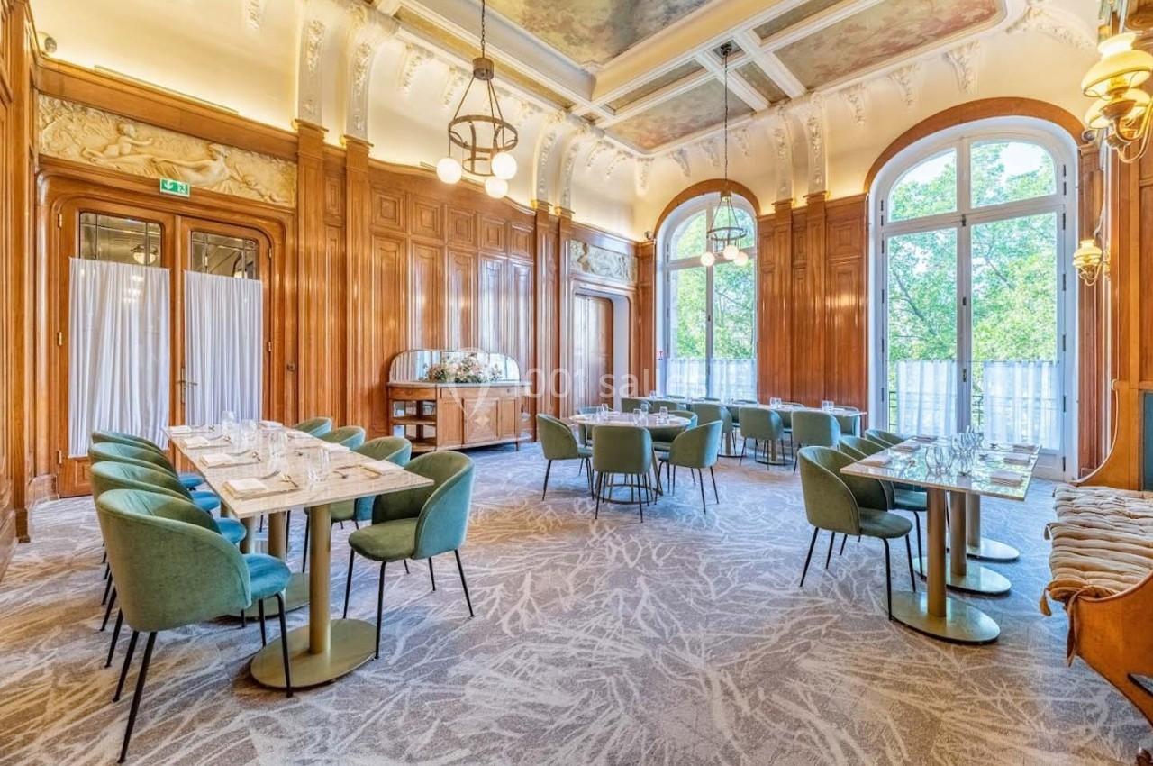 Salle élégante avec grandes fenêtres, murs en bois sculpté, plafond décoré et tables dressées avec chaises vertes.