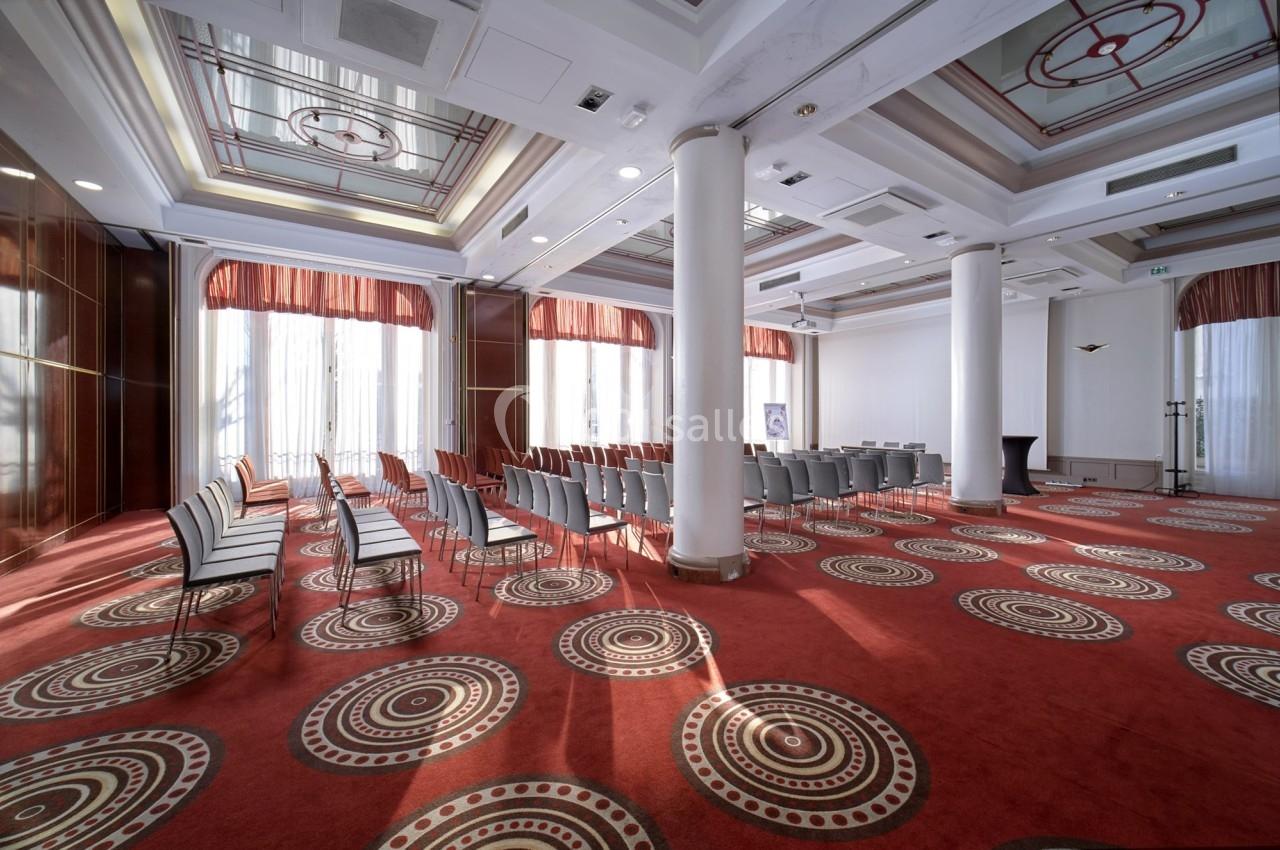 Salle de conférence lumineuse avec chaises alignées, tapis rouge à motifs circulaires et grandes fenêtres avec rideaux.