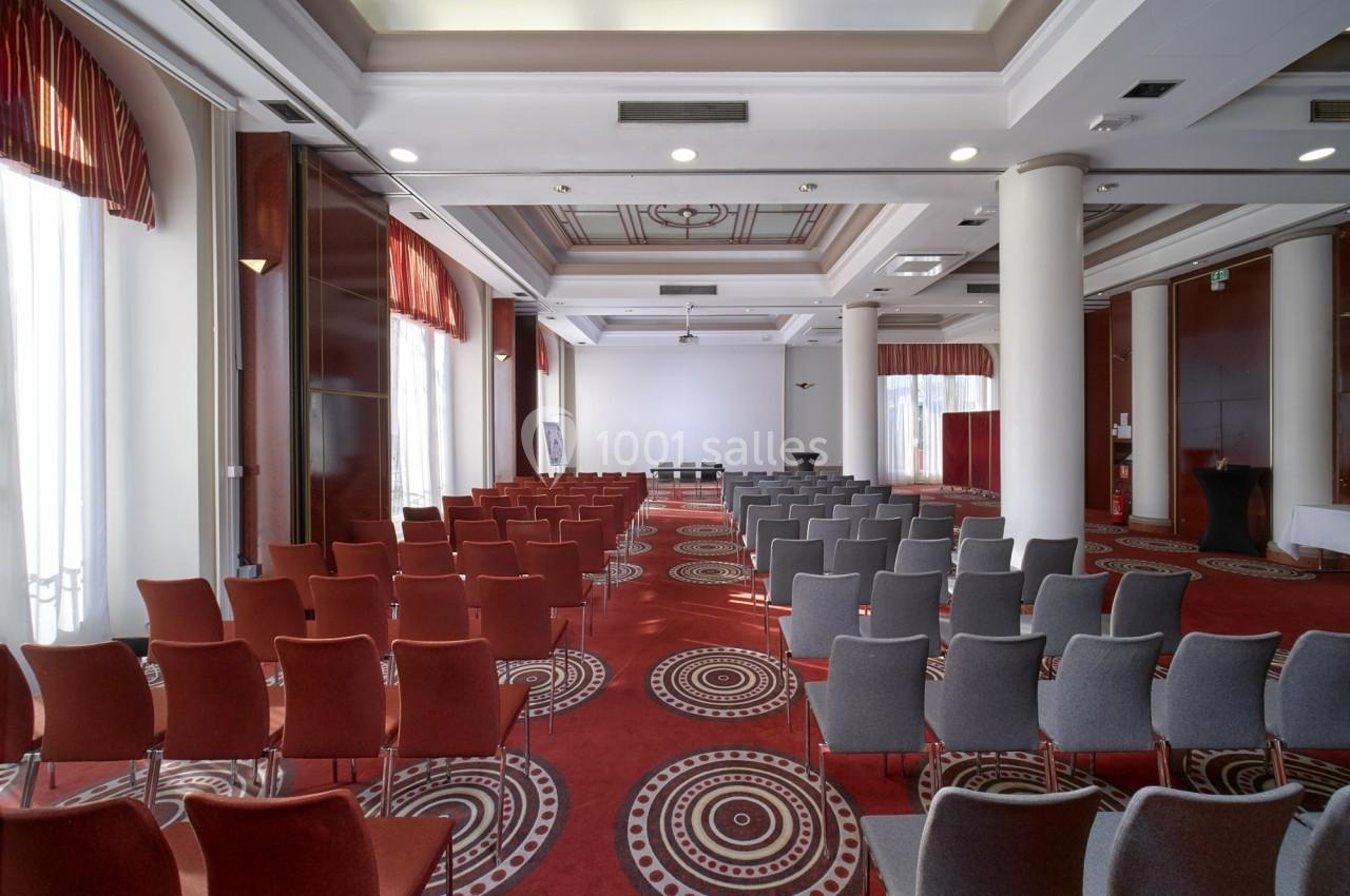 Salle de conférence avec rangées de chaises rouges et grises, tapis à motifs circulaires et lumière naturelle.