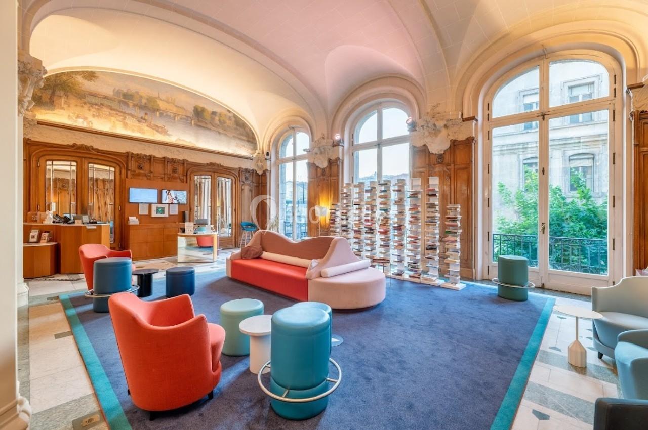 Salon lumineux avec canapés colorés, fauteuils modernes, étagères de livres et grandes fenêtres donnant sur l'extérieur.
