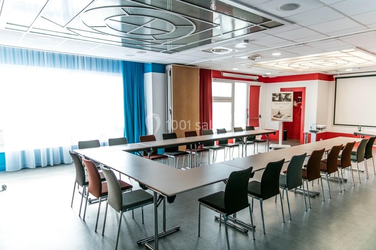 Salle de réunion lumineuse avec une grande table en U, des chaises colorées et un écran de projection.