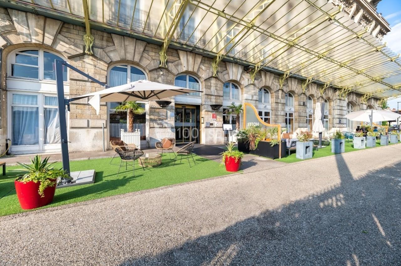 Terrasse extérieure avec tables, chaises et parasols devant un bâtiment en pierre sous une verrière.