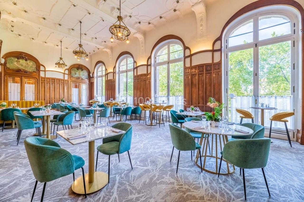 Salle de restaurant lumineuse avec grandes fenêtres, tables dressées et chaises vertes dans un décor élégant.