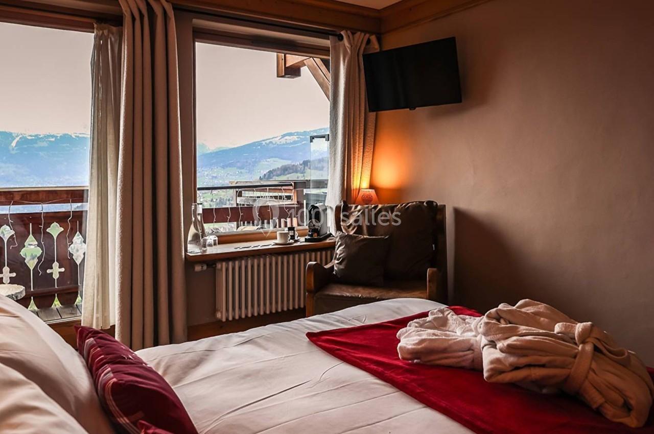 Chambre avec lit, peignoir posé, fauteuil, télé murale et vue sur des montagnes à travers une baie vitrée.
