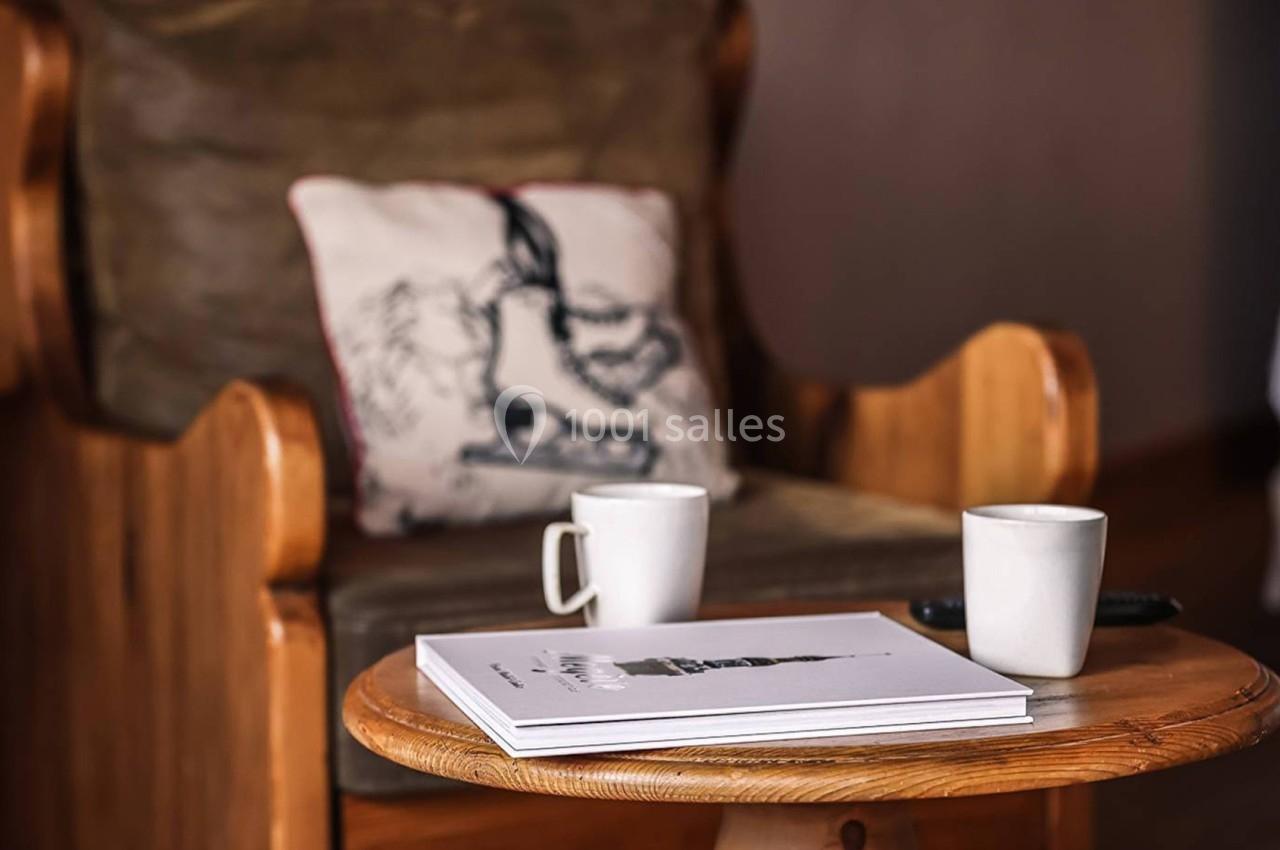 Deux tasses blanches et un magazine posés sur une table en bois devant un fauteuil avec un coussin illustré.