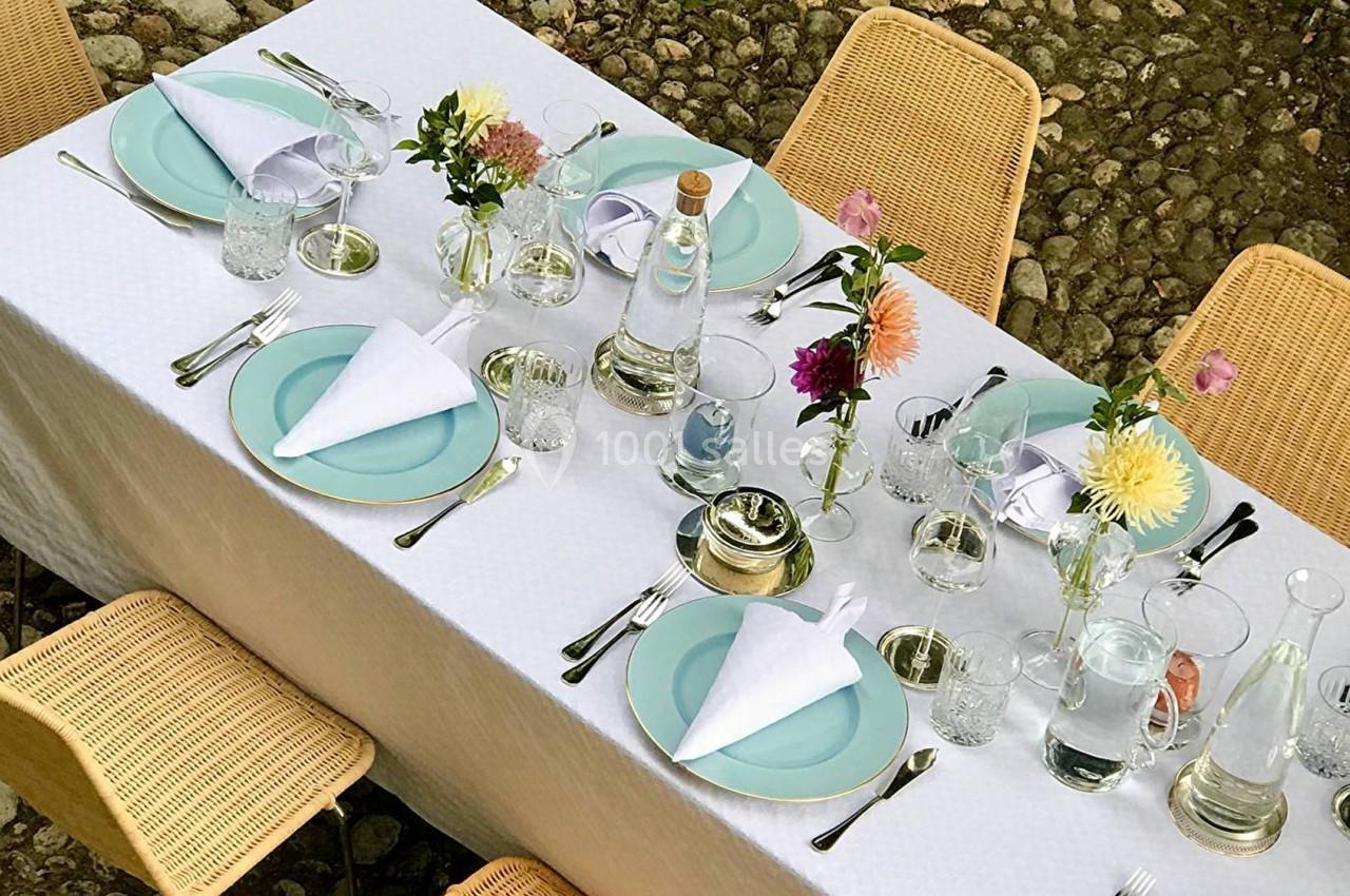 Table dressée en extérieur avec nappes blanches, assiettes pastel, verres, fleurs colorées et chaises en rotin.