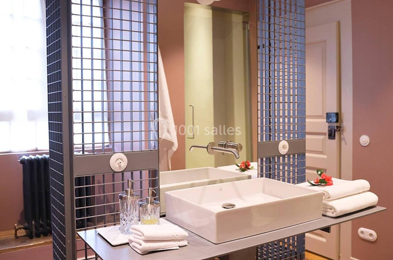 Lavabo moderne avec miroir grillagé, serviettes pliées et accessoires dans une salle de bain aux murs roses.