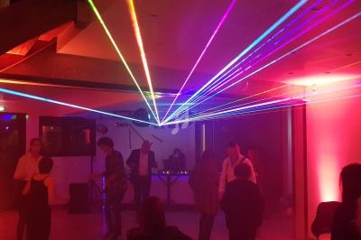 Personnes dansant dans une salle éclairée par des faisceaux laser colorés, avec un DJ en arrière-plan.