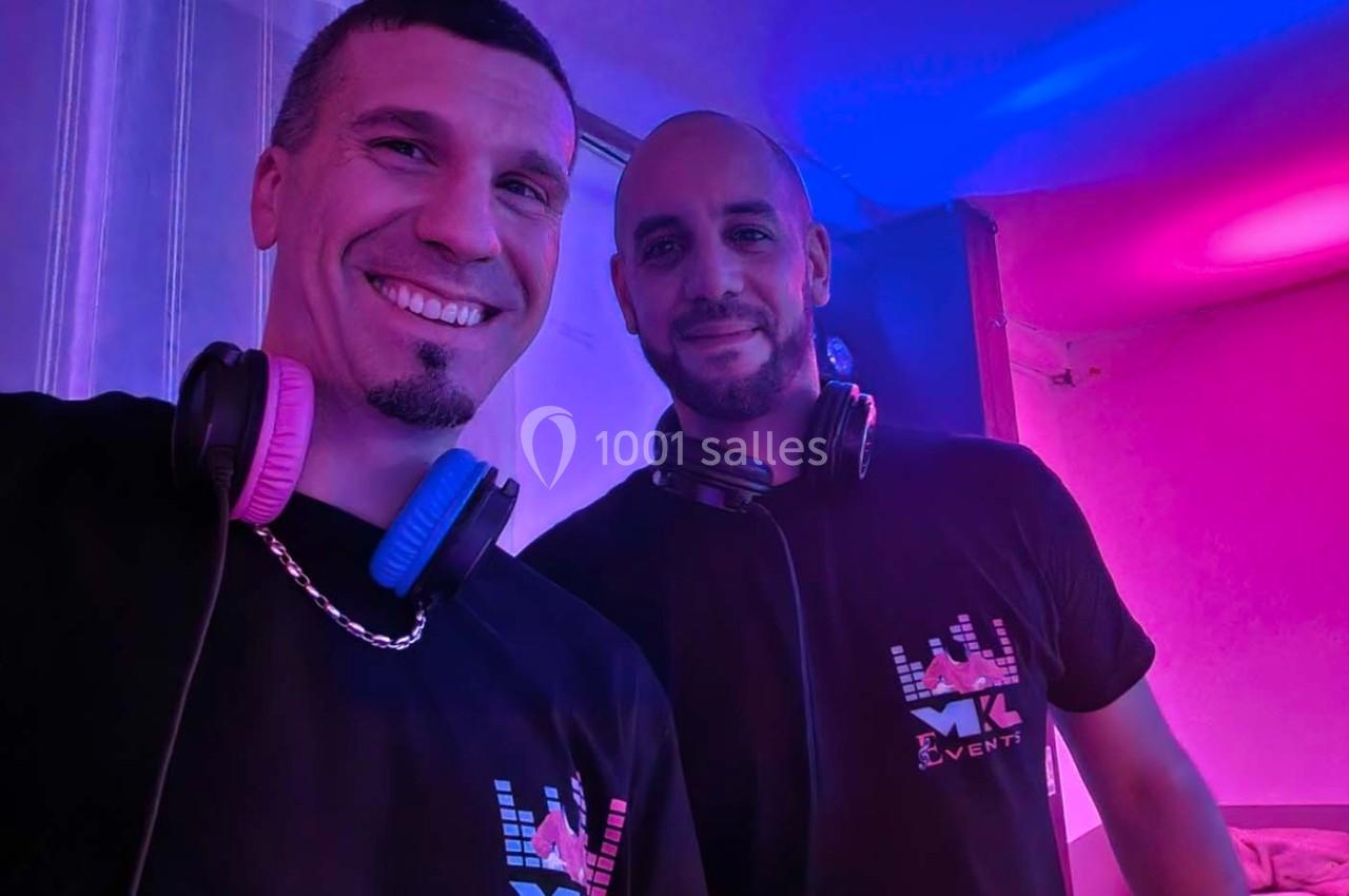 Deux hommes souriants portant des casques audio autour du cou, dans un environnement éclairé par des lumières colorées.