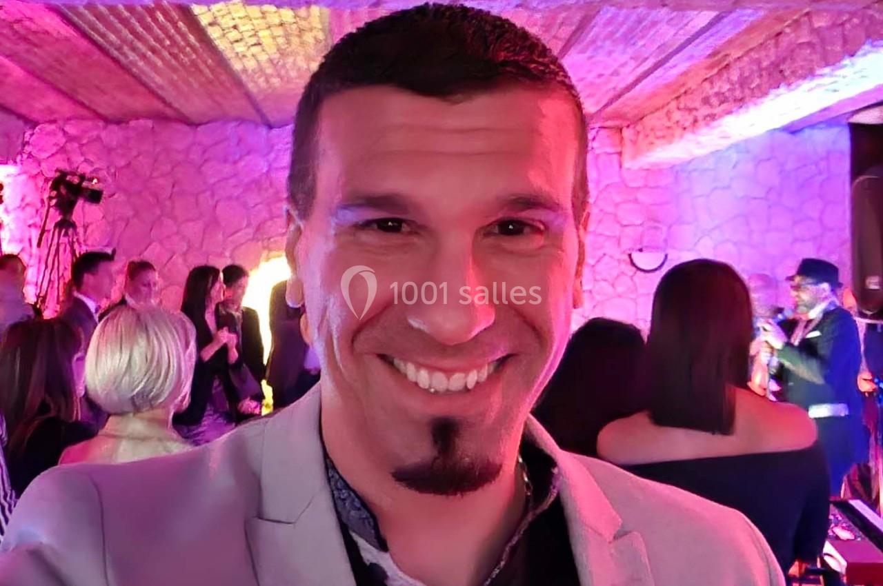 Homme souriant prenant un selfie lors d'un événement festif dans une salle éclairée aux tons roses et violets.