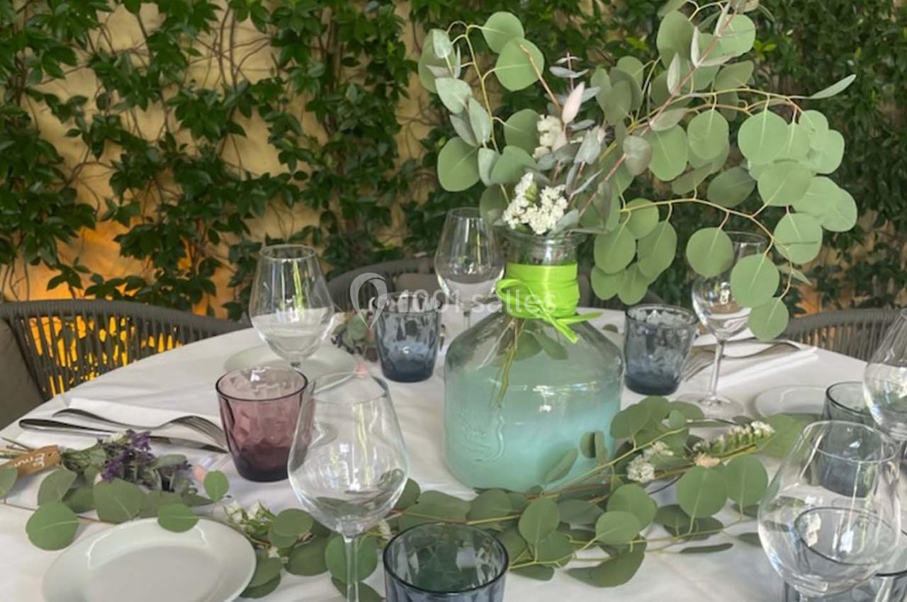 Table ronde décorée avec un bouquet de feuillage et de fleurs dans un vase en verre, entourée de vaisselle et de verres.