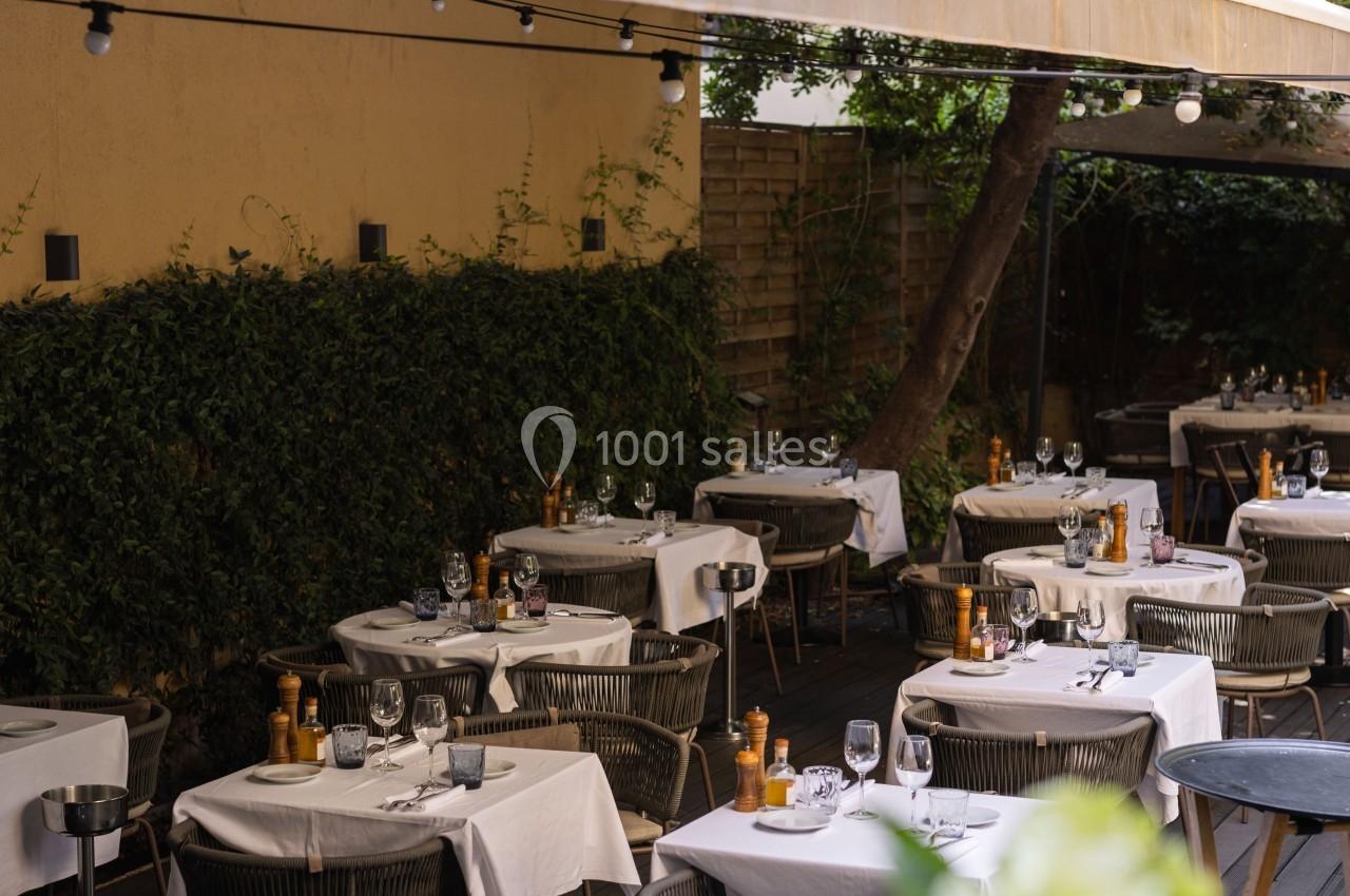 Terrasse de restaurant avec tables dressées, nappes blanches, chaises en osier et végétation en arrière-plan.