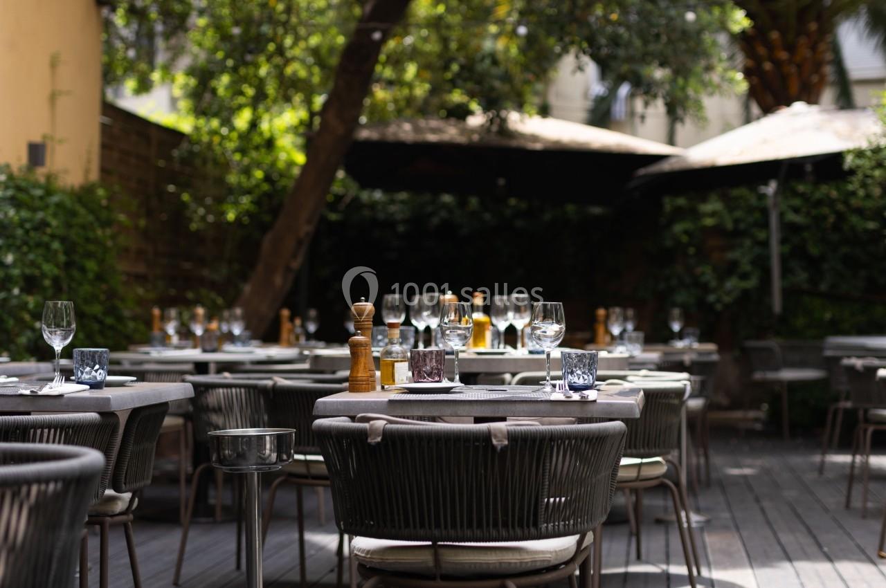 Terrasse d'un restaurant avec des tables dressées, chaises en rotin et parasols, entourée de verdure.