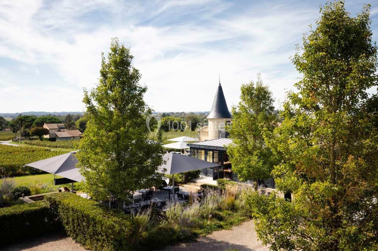Vue d'un domaine viticole avec terrasse ombragée, arbres, et une tour en arrière-plan sous un ciel dégagé.
