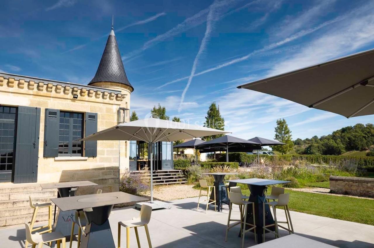 Terrasse extérieure d'un bâtiment en pierre avec parasols, tables et chaises, entourée de verdure sous un ciel bleu.