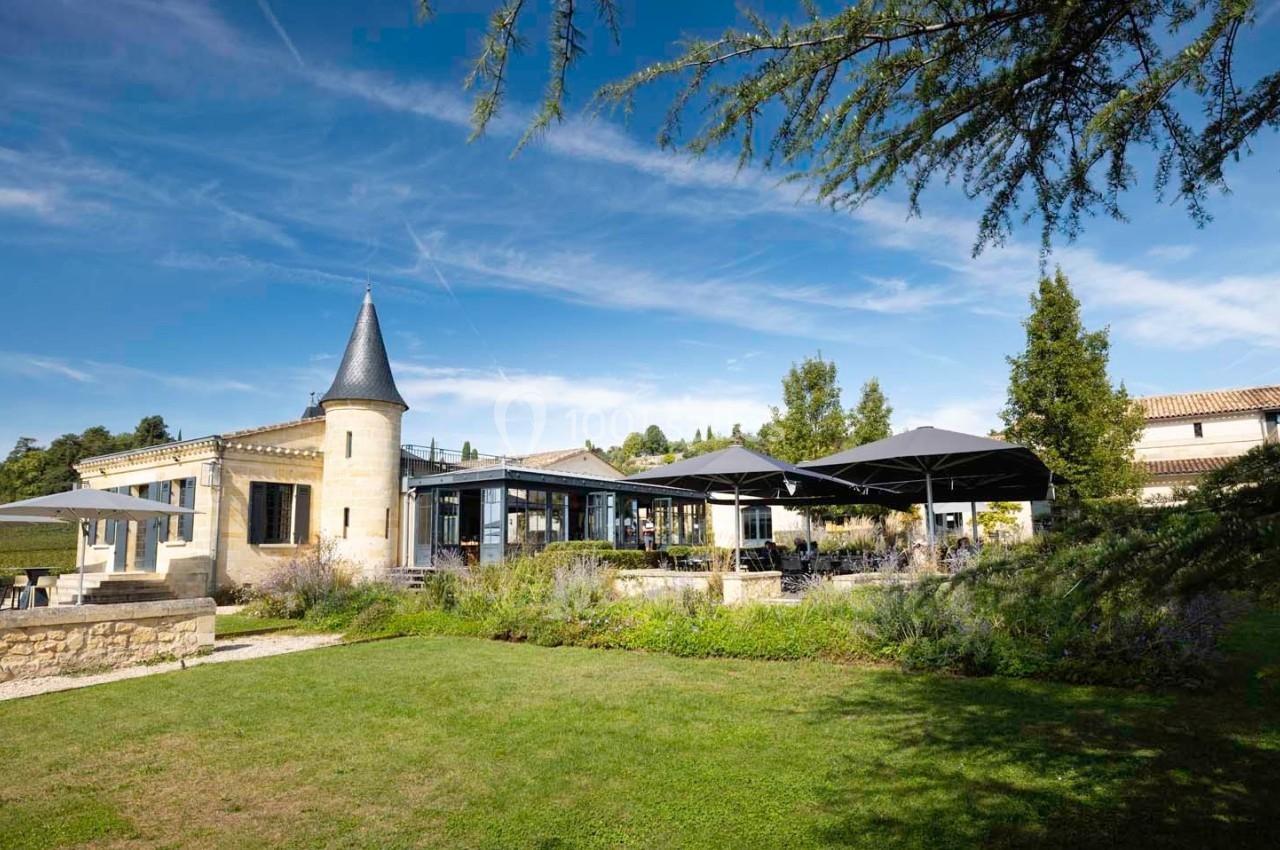 Bâtiment en pierre avec une tour, terrasse ombragée par des parasols, entouré de verdure sous un ciel bleu.