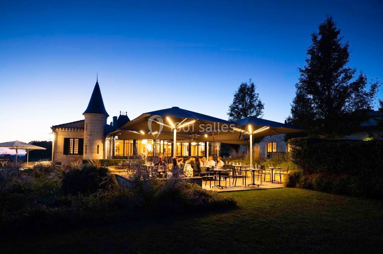 Terrasse éclairée d'un restaurant au crépuscule, avec des clients attablés et un bâtiment en pierre en arrière-plan.
