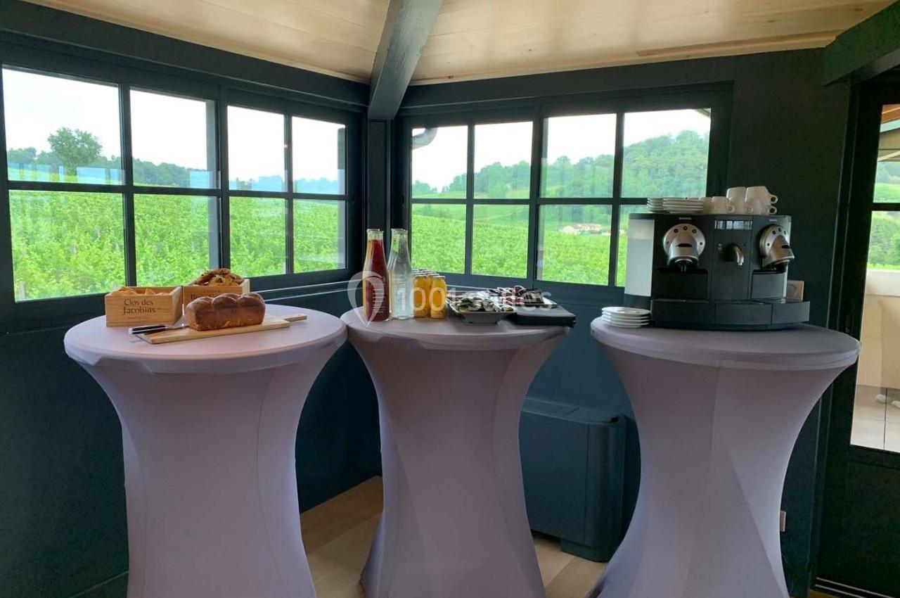 Trois tables hautes dressées avec boissons, viennoiseries et machine à café, dans une pièce lumineuse avec vue sur la…