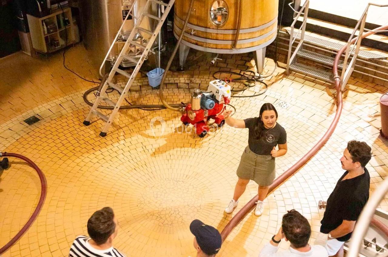 Une femme explique un processus à un groupe dans une brasserie, entourée d'équipements et de cuves en bois.