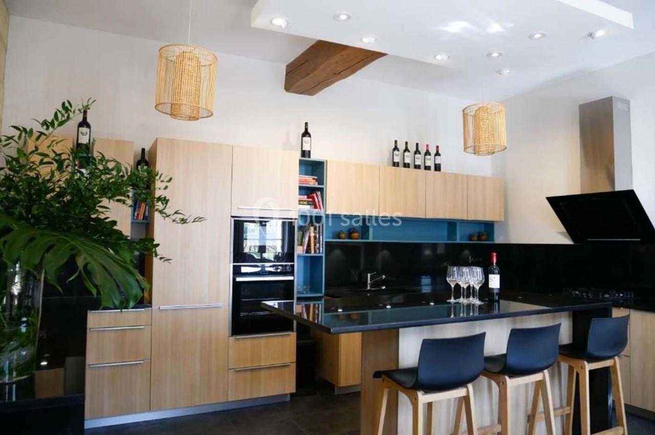 Cuisine moderne avec meubles en bois clair, plan de travail noir, tabourets hauts et éclairage suspendu.