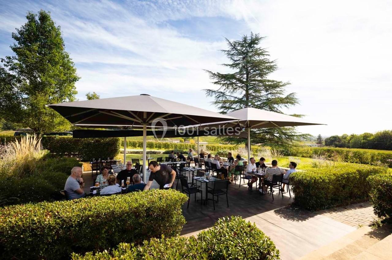 Terrasse extérieure d'un restaurant avec des clients assis sous de grands parasols, entourée de verdure.