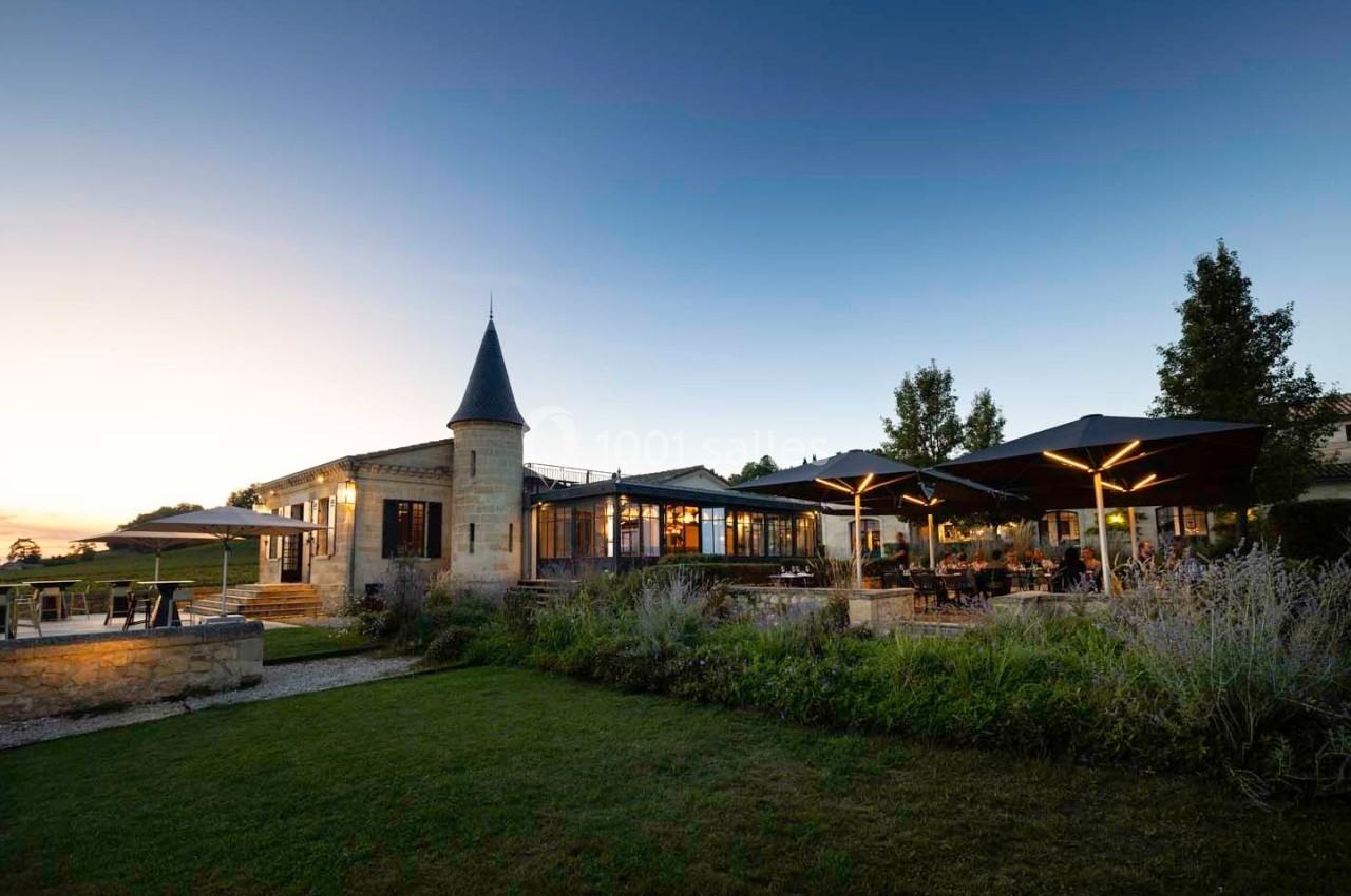 Bâtiment en pierre avec une tour, terrasse éclairée et parasols, entouré de verdure au crépuscule.