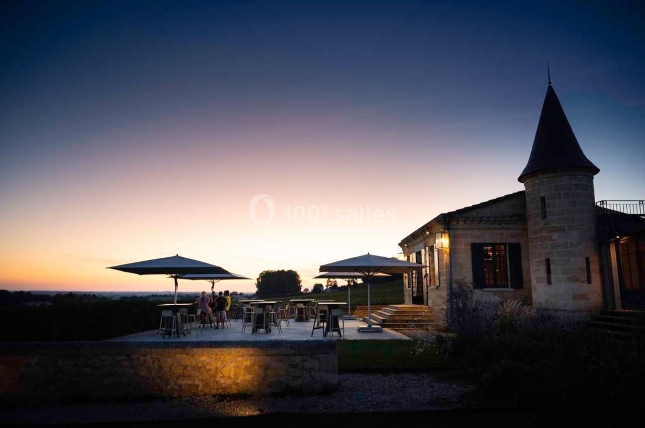 Terrasse éclairée d'un bâtiment en pierre avec parasols, au crépuscule, entourée de verdure.