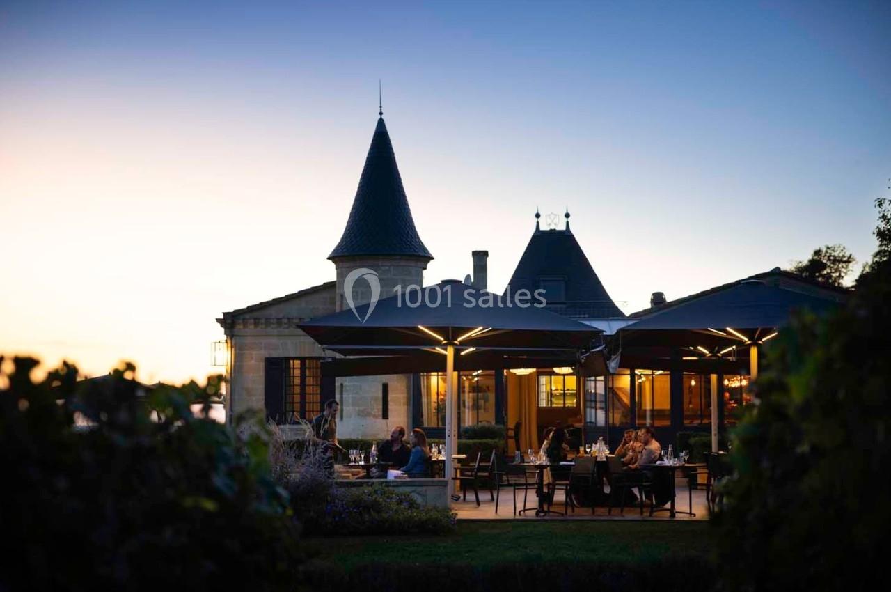 Terrasse éclairée d'un restaurant au crépuscule, avec des convives assis devant un bâtiment au style château.