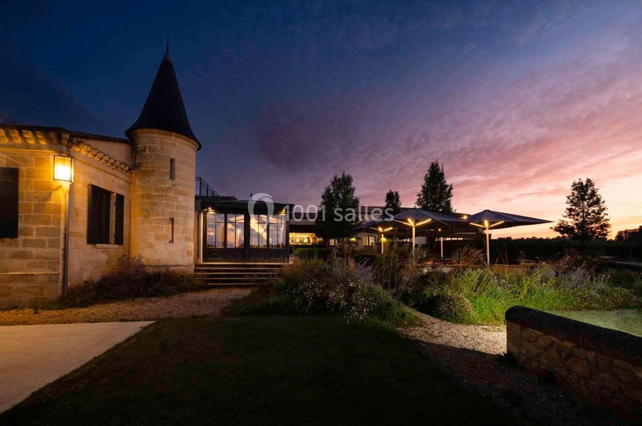 Bâtiment en pierre avec une tour, éclairé au crépuscule, entouré de végétation et de parasols illuminés.