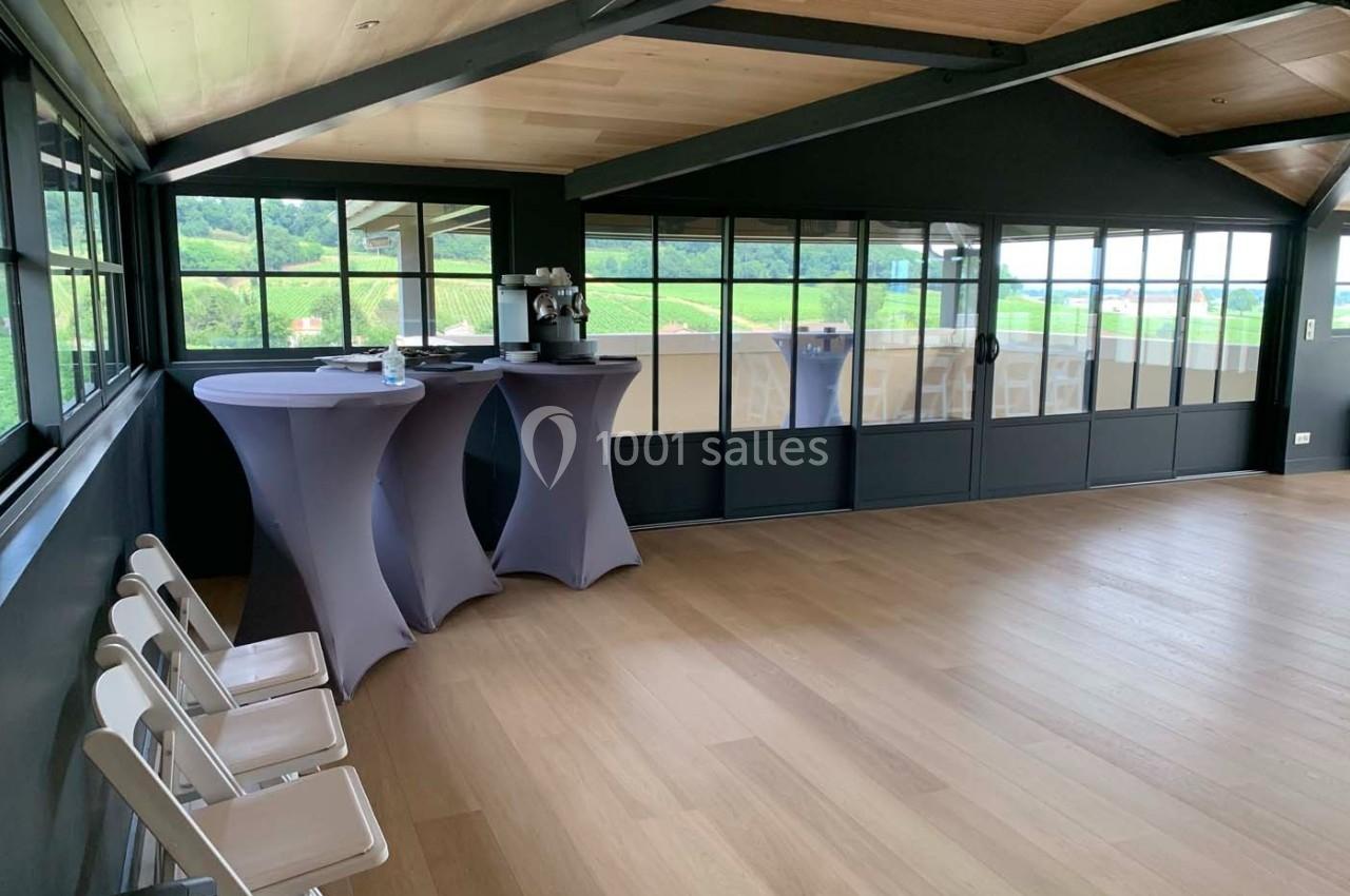 Salle lumineuse avec parquet en bois, grandes baies vitrées, tables hautes couvertes de nappes et chaises pliantes blanches.