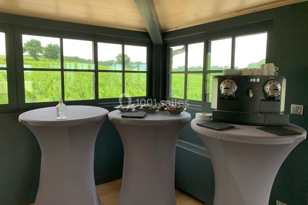 Trois tables hautes avec nappes violettes, une machine à café et des collations, dans une salle lumineuse avec vue sur la…