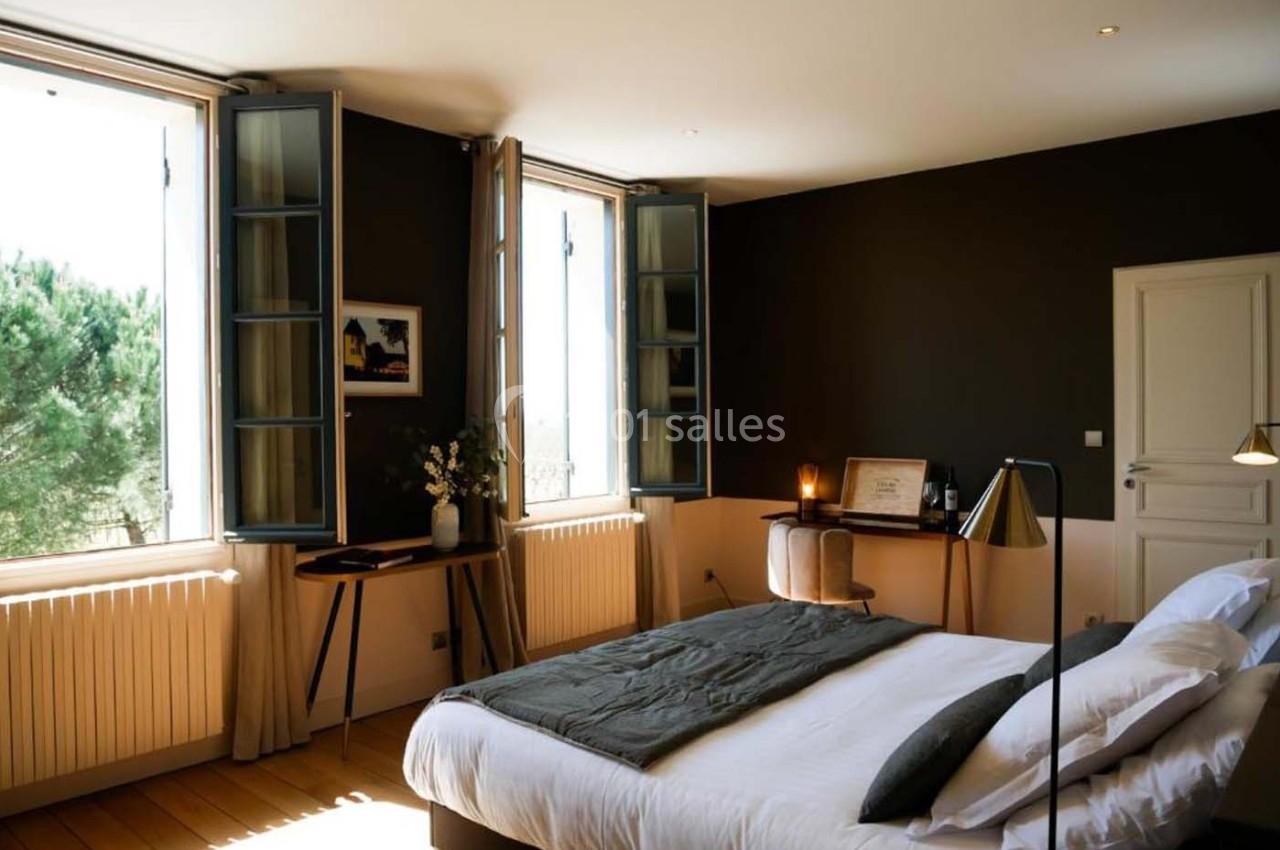 Chambre lumineuse avec lit double, bureau en bois, lampes et fenêtres ouvertes donnant sur des arbres.