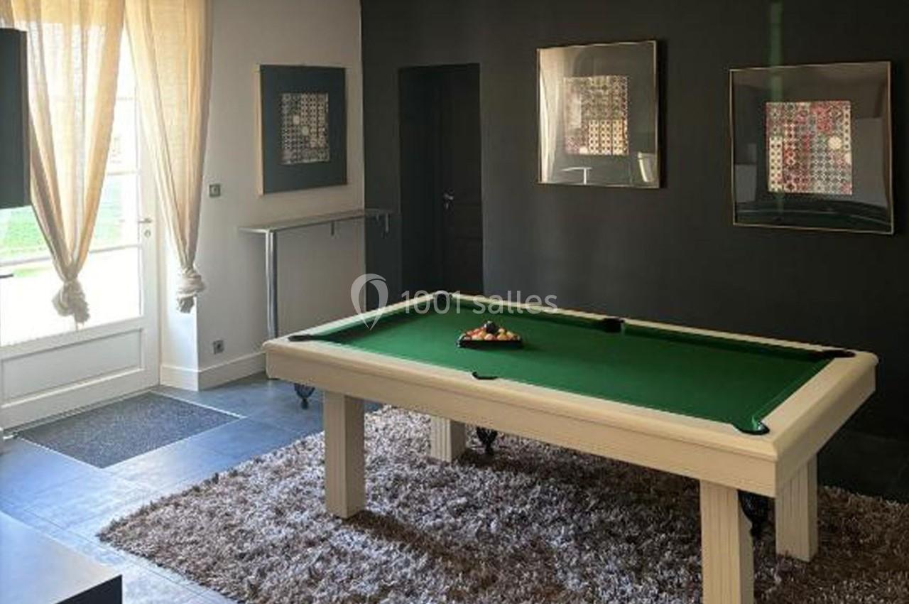 Table de billard blanche avec tapis vert, située dans une pièce sombre avec tapis épais et tableaux accrochés au mur.