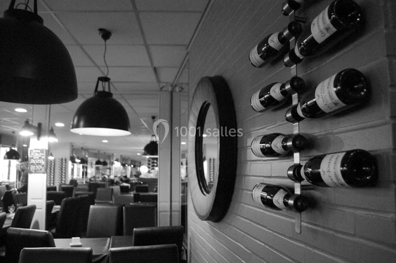 Mur avec des bouteilles de vin exposées, miroir rond et salle de restaurant avec chaises et suspensions noires en arrière…