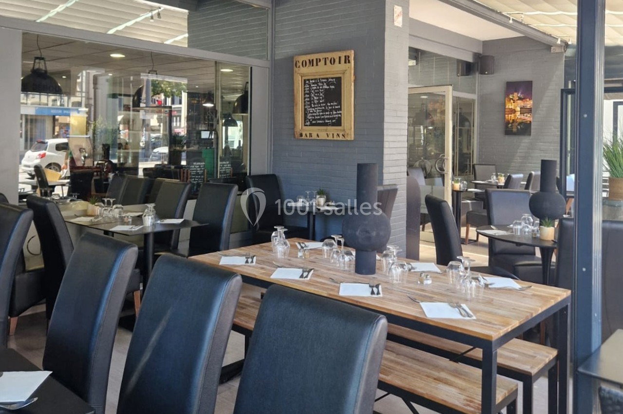 Salle de restaurant avec tables en bois, chaises noires, vaisselle dressée et vue sur l'extérieur vitré.