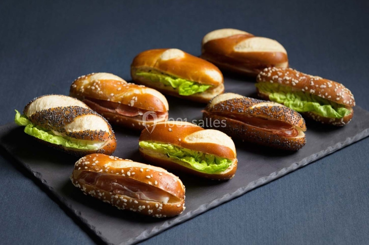 Assortiment de petits sandwichs sur pain bretzel garni de salade ou de jambon, disposés sur une ardoise noire.