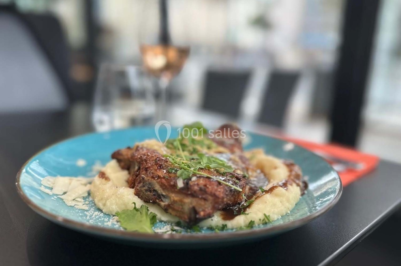 Plat de viande grillée sur purée, garni de verdure et copeaux de fromage, servi dans une assiette bleue.