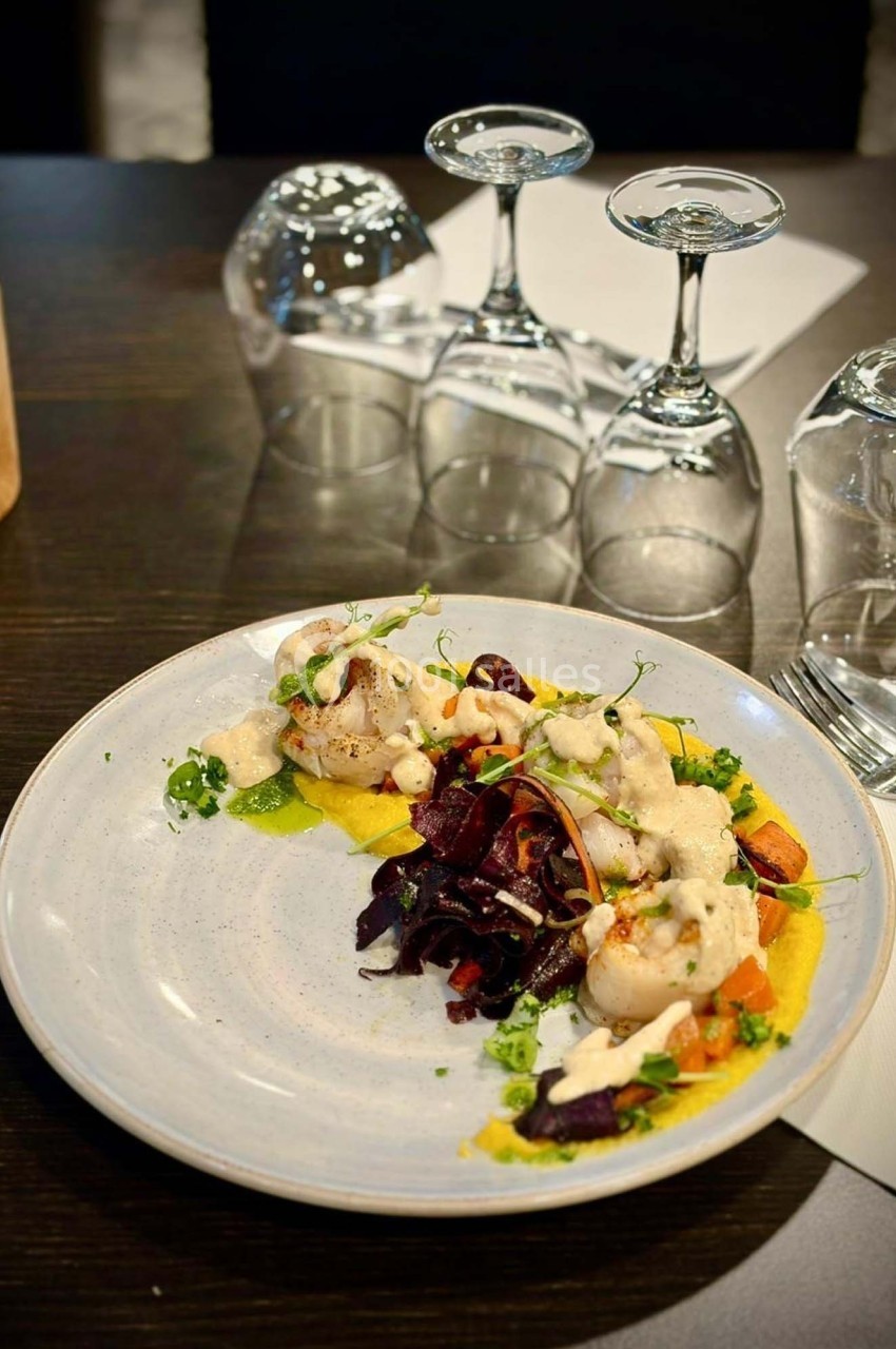 Assiette gastronomique avec légumes colorés, purée et sauce, servie sur une table avec verres à pied en arrière-plan.