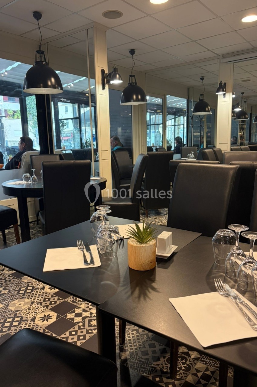 Salle de restaurant moderne avec tables dressées, chaises noires et grandes baies vitrées donnant sur l'extérieur.