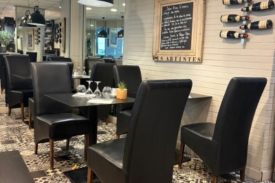 Table en bois dressée avec des sets noirs, des verres et des serviettes, située dans une véranda donnant sur une rue…