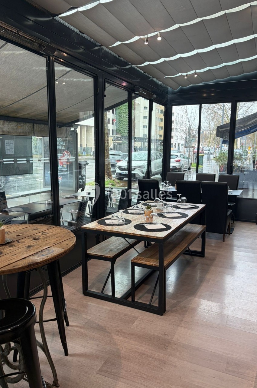 Salle de restaurant lumineuse avec tables en bois, chaises noires et baies vitrées donnant sur l'extérieur urbain.
