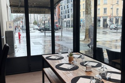 Table en bois dressée avec des sets noirs, des verres et des serviettes, située dans une véranda donnant sur une rue…